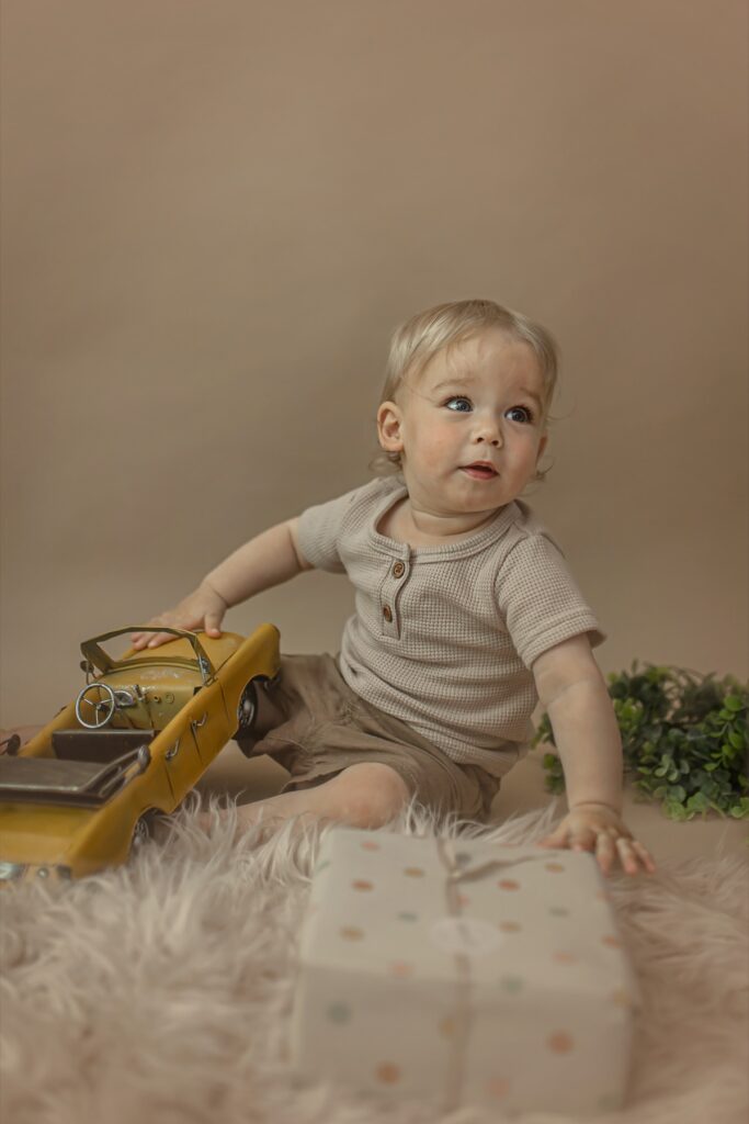 גיל שנה - פעוט יושב ברצפה עם מעטפה של מתנה הפעוט לובש מכנס וחולצה לבנה משחק עם מכונית צהובה